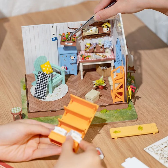 A DIY kit showcasing the Dreamy Garden House by Robotime. The image features a miniature interior with vibrant furniture, including a blue cabinet, a cozy green chair, and a rustic table set up with various decorations and plants. Hands are seen assembling a colorful orange shelf, highlighting the intricate details and craftsmanship of the model.