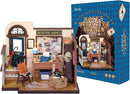 Mose's Detective Agency miniature model featuring a detailed office scene. The setup includes a wooden desk with a typewriter, a chair, a blue lamp, and a wall covered with investigation notes, maps, and photos. Accessories like a vintage telephone, a suitcase, and an umbrella stand are present, along with a decorative poster and a door leading outside.