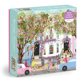 The Joy Laforme Spring Terrace jigsaw puzzle featuring a vibrant street scene adorned with blooming flowers and colorful houses. The puzzle box showcases a beautiful illustration with people walking, biking, and enjoying the spring atmosphere, perfect for anyone who loves puzzles and lively artwork.