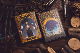 Two decks of King Star Magic Crystal Playing Cards displayed on a textured wooden surface. The cards feature intricate designs, with one deck showcasing a mysterious figure in a dark cloak holding a crystal, while the other deck portrays a vibrant blue cityscape against a starry background. Surrounding the cards are decorative elements like pinecones and wooden rounds, enhancing the enchanting theme.