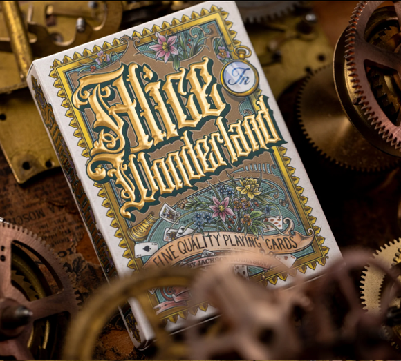 A close-up of the 'Alice in Wonderland' playing cards box, designed by Kings Wild. The box features intricate illustrations of flowers and whimsical designs, highlighted with golden lettering that reads 'Alice in Wonderland. Fine Quality Playing Cards.' It's beautifully displayed on a textured surface surrounded by mechanical gears.