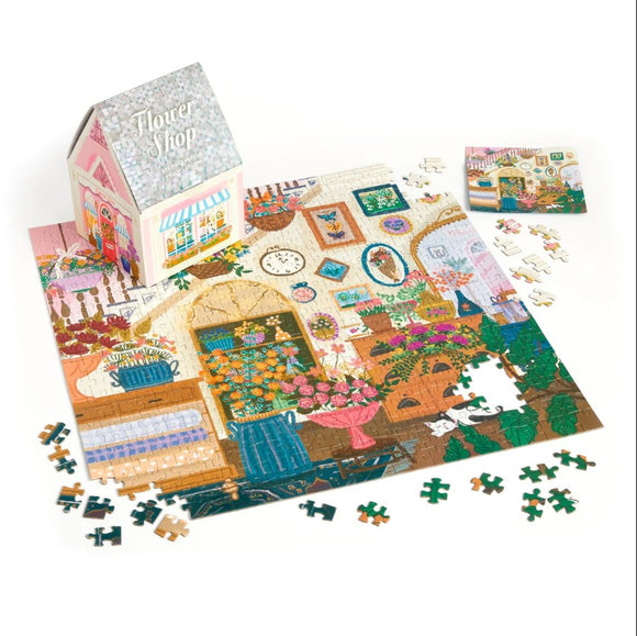 Joy Laforme Flower Shop jigsaw puzzle by Galison. The image shows a vibrant floral-themed puzzle depicting a charming flower shop setup. The puzzle pieces are scattered around the nearly completed puzzle, which features colorful flowers, potted plants, and decorative elements within a cozy flower shop setting. A whimsical puzzle box resembling a flower shop is positioned in the background.