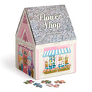Joy Laforme Flower Shop jigsaw puzzle by Galison. The puzzle features a charming flower shop with a pink exterior, colorful flower displays, and a welcoming 'Open' sign. The packaging is designed to resemble a house with a shingled roof, and several puzzle pieces are scattered in front.