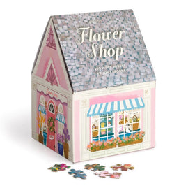 Joy Laforme Flower Shop jigsaw puzzle by Galison. The puzzle features a charming flower shop with a pink exterior, colorful flower displays, and a welcoming 'Open' sign. The packaging is designed to resemble a house with a shingled roof, and several puzzle pieces are scattered in front.