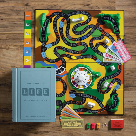 An overhead view of The Game of Life Vintage Books game set up on a wooden surface. The colorful game board features a winding path with spaces labeled for various life events. A vintage-looking book titled 'The Game of Life' is placed on the side, with a wheel spinner and various money cards, houses, and game pieces scattered around, showcasing the complete gameplay experience.