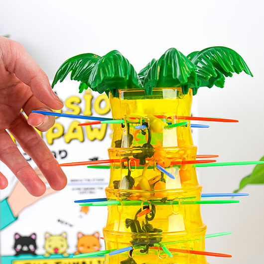 A hand interacts with the 'Mission Impawsible' game by Gift Republic, featuring a colorful toy structure resembling a palm tree. The game includes various colored sticks and small animal figures inside, designed for a fun and exciting challenge where players must carefully remove the sticks without letting the animals fall.