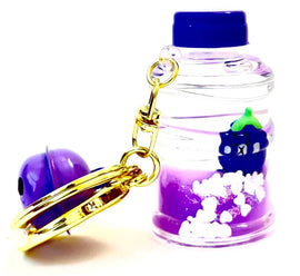 A colorful Eggplant Floaty Keychain from BC Mini featuring a clear bottle containing purple liquid and glitter, with a small green-topped eggplant figure inside. The keychain includes a shiny golden clasp and a purple round charm, creating a whimsical accessory perfect for bags or keys.