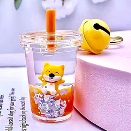 A colorful Shiba Inu floaty keychain displayed in a clear cup filled with water, featuring a cheerful yellow Shiba Inu figurine inside, surrounded by colorful glitter. An orange straw emerges from the cup, and a small yellow bell keychain is visible in the background, adding a playful touch.