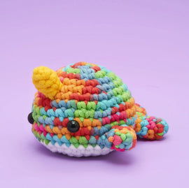 A colorful Rainbow Narwhal crochet kit featuring vibrant yarn in various shades of red, yellow, green, blue, and pink. The narwhal has a distinct yellow horn and large black eyes, set against a soft purple background, showcasing its adorable design perfect for kids and beginners. Ideal for DIY enthusiasts looking to create their own plush toy.