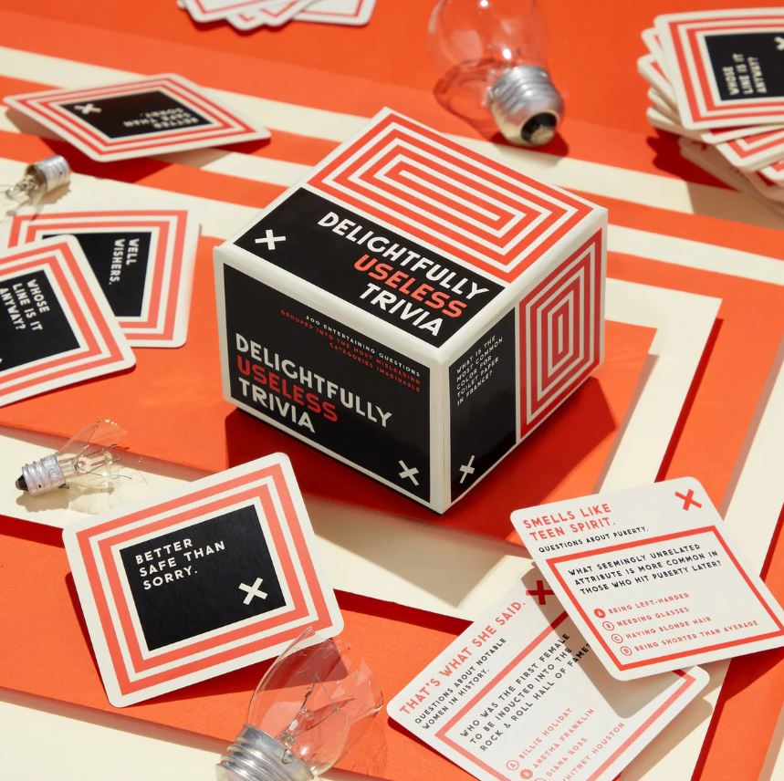 A vibrant display of the 'Delightfully Useless Trivia' game by Brass Monkey. The cube-shaped box features bold red and black graphics, with trivia question cards scattered around. Several light bulb props add a fun touch, visually enhancing the playful theme of trivia and knowledge. Perfect for game nights with friends or family, this game promises laughter and entertainment with its quirky questions.