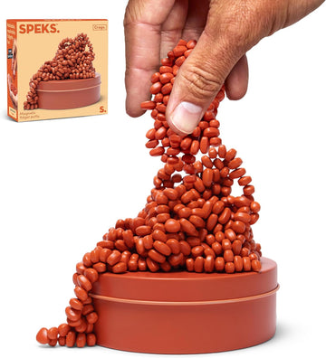 A hand holds a cluster of coral-colored magnetic fidget putty from the Crags collection. The putty is shown in a dynamic form, resembling a figure, arranged on a round, matching base. The product packaging appears in the top left, showcasing its playful design and vibrant colors.