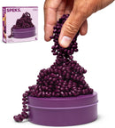A hand is reaching for a pile of purple magnetic fidget putty beads that are cascading from a round storage container. The container is purple and labeled 'SPEKS Crags Matte: Iris.' The putty is shown in a whimsical, flowing arrangement, inviting tactile exploration.