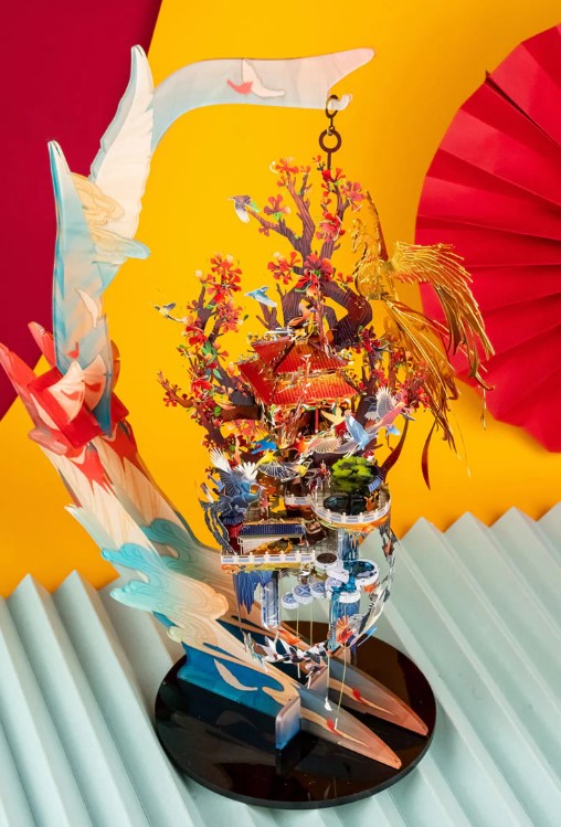 The Flaming Plume Stand for Metal Model by Mu Model features a captivating and colorful design, showcasing a symphony of vibrant flowers, a majestic phoenix, and intricate architectural elements. Set against a bright yellow backdrop, the stand appears dynamic and artistic, ideal for displaying metal models.