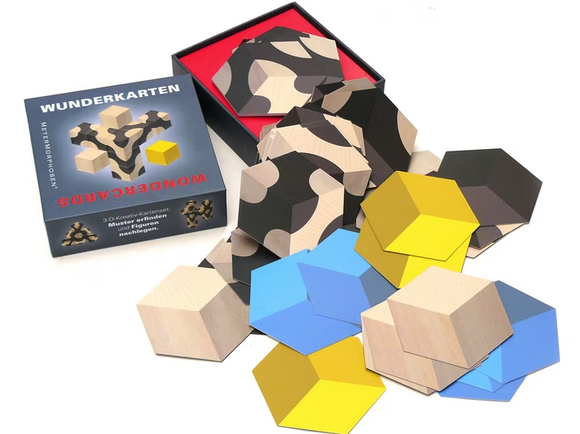 The Wunderkarten - Illusions mechanical puzzle set by MeterMorphosen. It features a colorful array of hexagonal wooden pieces in blue, yellow, and black patterns, scattered around an open black box with a red interior. The pieces are designed for intricate arrangement, promoting creative problem-solving.