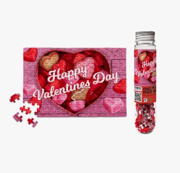 A colorful jigsaw puzzle titled 'Happy Valentine's Day' featuring a heart shape filled with chocolate and heart-shaped candies. The puzzle is partially assembled and surrounded by a few scattered puzzle pieces. It comes in a transparent container showcasing small puzzle pieces.