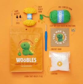 Fred the Dinosaur Beginner Crochet Kit by The Woobles. The kit includes easy peasy yarn, pre-started yarn, safety eyes, stuffing, a crochet hook, and a yarn needle, all packaged in a vibrant orange bag featuring a cute dinosaur design. Step-by-step video instructions are also included to guide beginners in crocheting their own dinosaur.