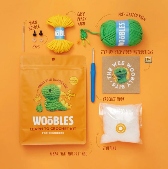 Fred the Dinosaur Beginner Crochet Kit by The Woobles. The kit includes easy peasy yarn, pre-started yarn, safety eyes, stuffing, a crochet hook, and a yarn needle, all packaged in a vibrant orange bag featuring a cute dinosaur design. Step-by-step video instructions are also included to guide beginners in crocheting their own dinosaur.