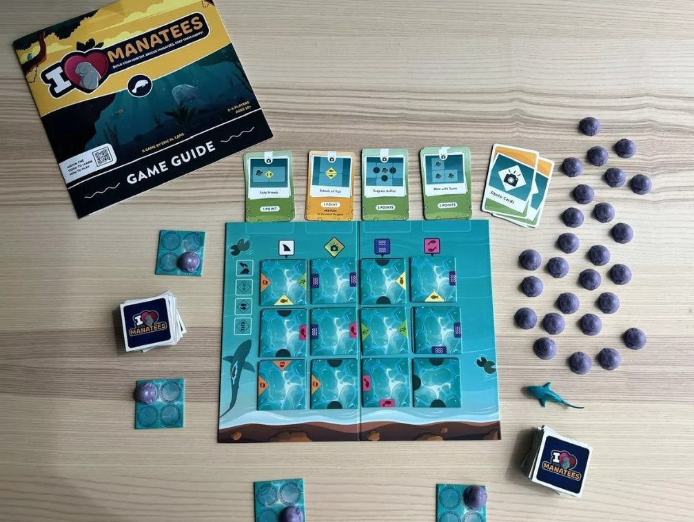 An organized game layout of the 'I Heart Manatees' game by Buffalo Games, featuring a game board with tiles and various tokens, a game guide on the top left, and action cards laid out showing point values. The colorful design includes water patterns, manatee illustrations, and player tokens, all arranged on a wooden table.