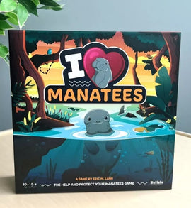 The I Heart Manatees game box by Buffalo Games features vibrant artwork of manatees in a serene water setting surrounded by lush trees. The title is prominently displayed with a heart symbol, emphasizing the theme of love for manatees. Suitable for ages 10 and up, the game supports 2 to 4 players.