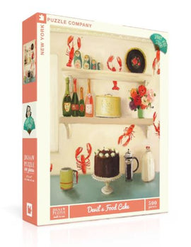 The packaging of the Devil's Food Cake jigsaw puzzle by New York Puzzle Company, featuring a vintage-style kitchen scene with a chocolate cake centerpiece, vibrant flowers, lobster motifs, and various bottles and jars on shelves. The puzzle contains 500 pieces and is designed for both adults and children.