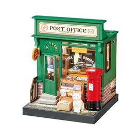 A detailed miniature model of a green post office building featuring a door, large window, and red post box. The model includes decorative elements such as a sign and a small white cat on the steps, representing a charming DIY kit for crafting enthusiasts.