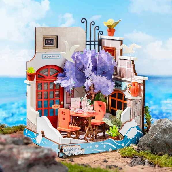 A beautifully detailed DIY kit of the Sea Holiday Restaurant, showcasing a charming eatery by the ocean. The model features vibrant colors, a purple tree, outdoor seating with orange chairs, decorative plants, and whimsical details such as birds and a wave motif. Perfect for hobbyists, this kit creates a serene seaside ambiance.