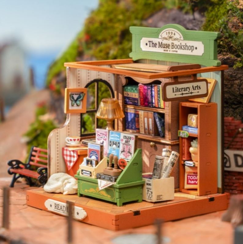 A detailed miniature model of 'The Muse Bookshop,' showcasing intricately designed shelves filled with books, a cozy lamp, and a vibrant storefront sign. The model features decorative items, including a green storage box and a small bench outside, emphasizing the quaint, inviting atmosphere of a literary-themed shop.