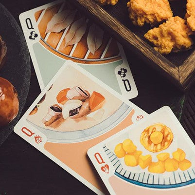 A close-up of Night Market: Day Playing Cards featuring beautifully illustrated cards depicting various delicious food items. The cards are spread on a dark wooden surface with some fried chicken and dessert in the background, highlighting the vibrant colors and intricate designs of the food illustrations on the cards.