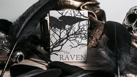 The Raven Black Dusk Playing Cards by WJPC, featuring a striking design with silhouettes of a raven amid intricate branches against a stark white background. The card box is surrounded by dark feathers and a skull, enhancing its mysterious and gothic theme.