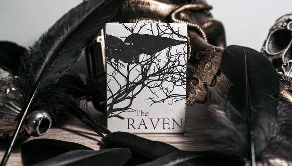 The Raven Black Dusk Playing Cards by WJPC, featuring a striking design with silhouettes of a raven amid intricate branches against a stark white background. The card box is surrounded by dark feathers and a skull, enhancing its mysterious and gothic theme.