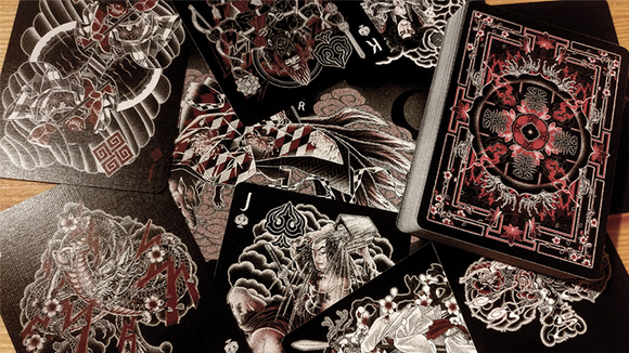 A close-up view of Sumi Original Craft Playing Cards spread out, showcasing intricate black and white artwork with hints of red. The deck features unique designs on each playing card, featuring mythical and traditional motifs, laid beside the embossed card box.