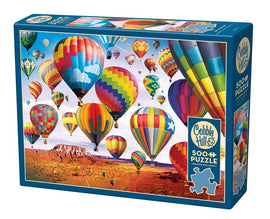 A Cobble Hill jigsaw puzzle titled 'Up in the Air' featuring a colorful scene of hot air balloons soaring above a desert landscape. The box contains 500+ pieces and includes a poster for reference. The vibrant balloons come in various patterns and colors, creating a cheerful and whimsical atmosphere.