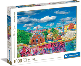 Clementoni 1000-piece jigsaw puzzle featuring the vibrant and colorful landscape of Park Güell in Barcelona. The box displays a scenic view of the architectural wonder, showcasing the intricate mosaic details and lush greenery under a bright blue sky with fluffy clouds, perfect for puzzle enthusiasts and fans of Gaudí's work.
