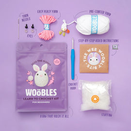 Jojo the Bunny Beginner Crochet kit by Woobles, featuring a colorful packaging with a cute bunny design. The kit includes easy peasy yarn, a crochet hook, yarn needle, eyes for the bunny, pre-started yarn, stuffing, and step-by-step video instructions, all neatly arranged on a purple background.