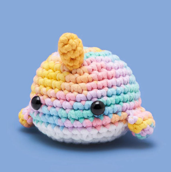 A cute pastel narwhal plush toy made of colorful crocheted yarn. The narwhal features a spiral horn and large black button eyes, with a soft gradient of pink, blue, yellow, and white colors. It sits on a light blue background, ideal for beginners in crocheting.