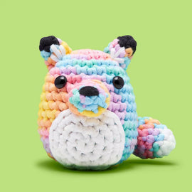 A colorful pastel fox made from crochet yarn, featuring a round body with vibrant rainbow colors and a white belly. The fox has large, shiny black eyes and small ears, sitting against a green background, ideal for beginner crochet enthusiasts.