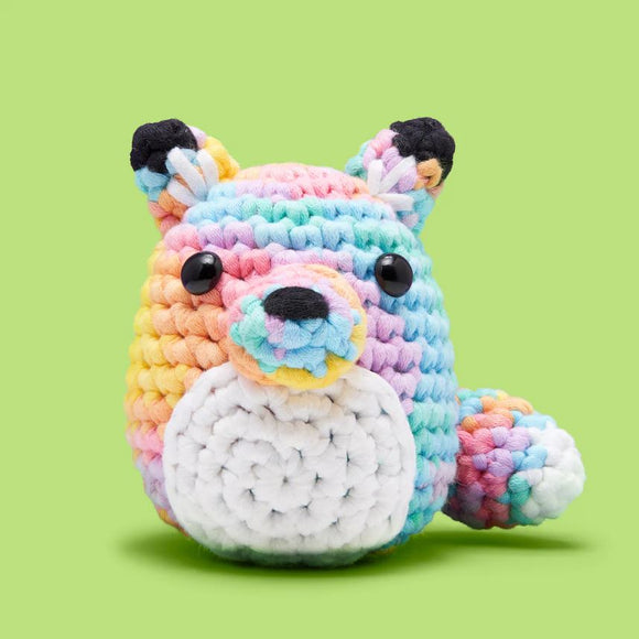 A colorful pastel fox made from crochet yarn, featuring a round body with vibrant rainbow colors and a white belly. The fox has large, shiny black eyes and small ears, sitting against a green background, ideal for beginner crochet enthusiasts.