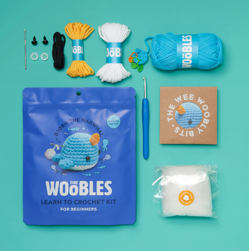 Bjorn the Narwhal Beginner Crochet Kit by Woobles, featuring a blue packaging with a cute narwhal design. The kit includes various yarns in blue, yellow, white, and black, safety eyes, and a crochet hook, all arranged on a teal background.