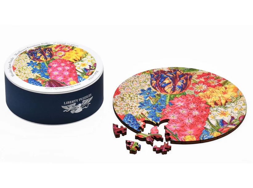 The Mayflower Premium Bulbs jigsaw puzzle by Liberty Puzzles is presented in an elegant round box with a vibrant floral design on the lid. The puzzle pieces are displayed beside the box, showcasing intricate flower patterns in bright colors, including tulips and other blossoms, inviting puzzle enthusiasts to create a beautiful floral scene.