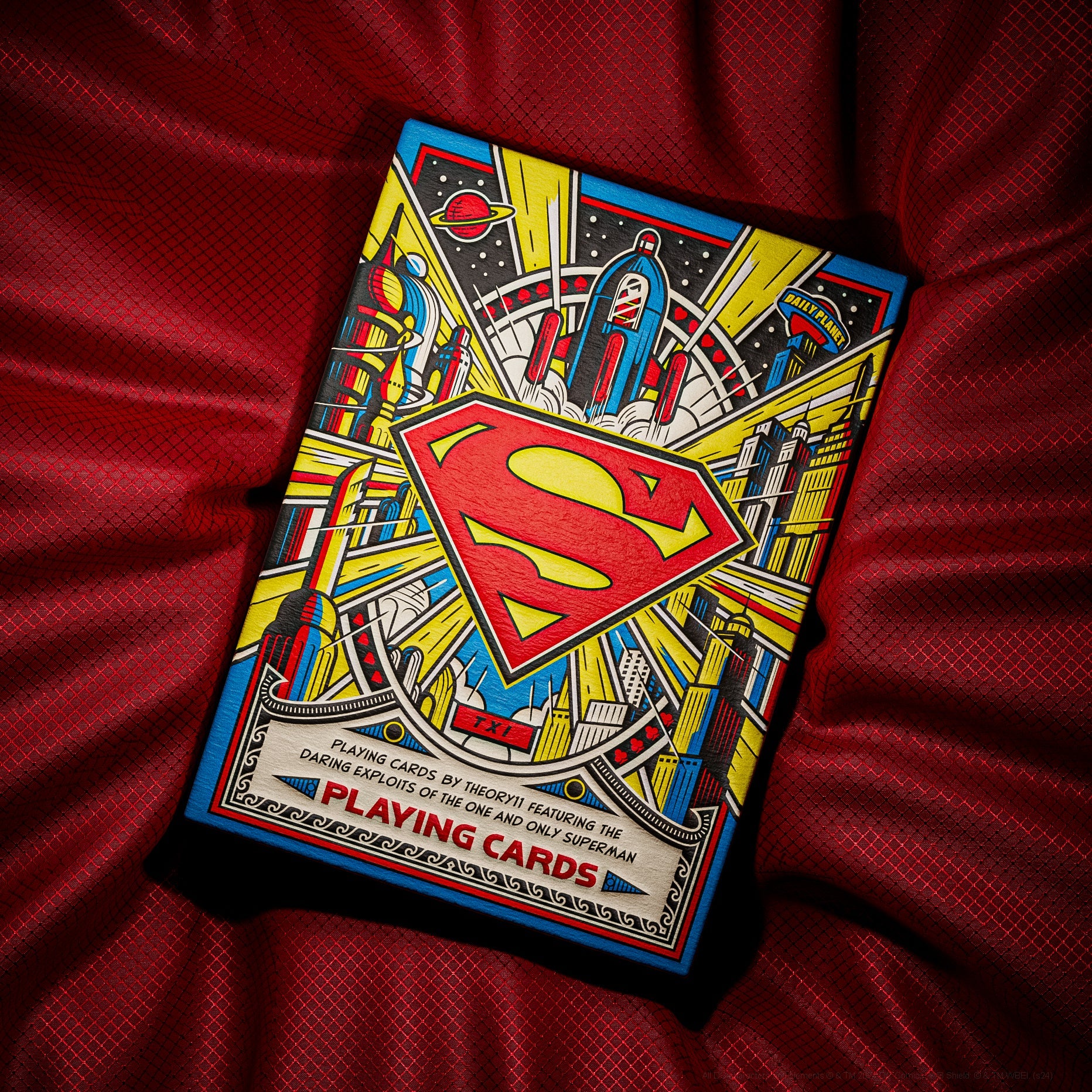 Superman Card Deck by Theory 11 displayed on a red fabric background. The deck features vibrant artwork showcasing Superman amidst a dynamic comic-style design, with elements representing space and cityscapes. The packaging includes bold colors and iconic imagery of Superman's emblem, making it a perfect addition for fans and collectors.