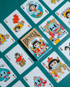 A vibrant display of Paper Royals Playing Cards by RunIt Decks, showcasing an array of colorful and whimsical illustrated cards. The deck is centered in the image, featuring a playful design with cartoon characters representing royalty. Surrounding the deck are several face cards, each adorned with unique illustrations that highlight a whimsical theme, set against a teal background.