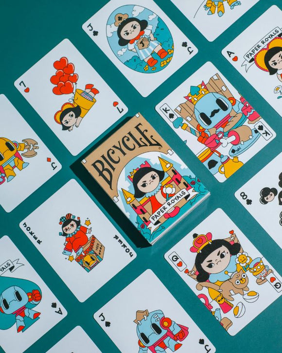 A vibrant display of Paper Royals Playing Cards by RunIt Decks, showcasing an array of colorful and whimsical illustrated cards. The deck is centered in the image, featuring a playful design with cartoon characters representing royalty. Surrounding the deck are several face cards, each adorned with unique illustrations that highlight a whimsical theme, set against a teal background.
