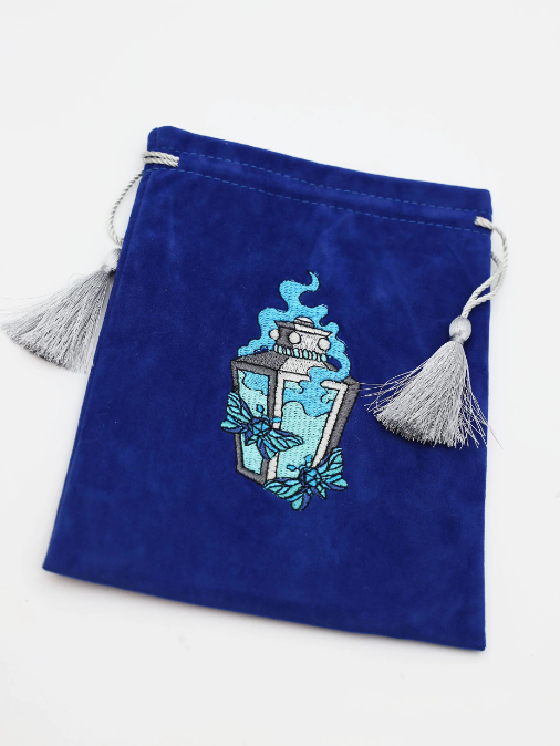 A plush blue dice bag featuring an intricately embroidered lantern design in shades of blue and gray. The bag is accented with silver tassels on both sides, perfect for storing gaming dice.