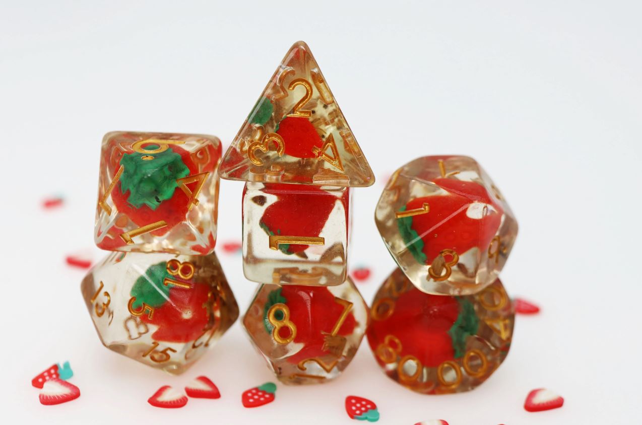 A set of Wild Strawberries RPG dice featuring vibrant red strawberries encased in clear resin. The dice showcase various shapes including a d20, d12, d10, d8, and d4, all displaying gold numbers for easy reading. Scattered around the dice are small strawberry-shaped confetti pieces, adding a playful touch to the display.