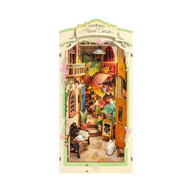 A beautifully detailed Floral Corner Book Nook DIY kit by Robotime showcases a charming miniature scene featuring a cozy café with flowers, plants, and a small white cat. The intricate design captures a whimsical ambiance perfect for book lovers and DIY enthusiasts.