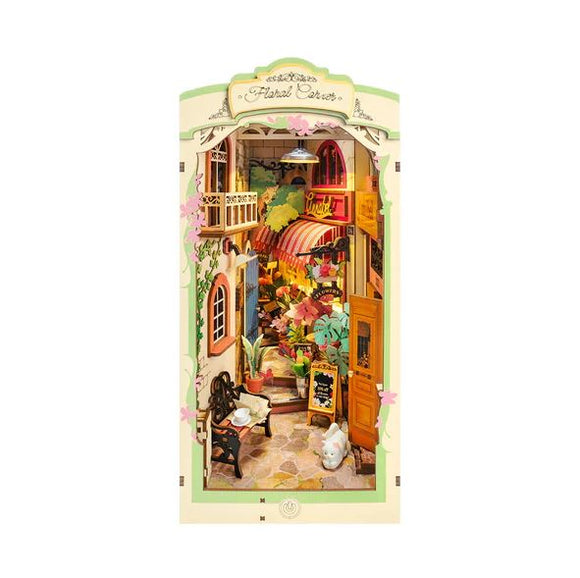 A beautifully detailed Floral Corner Book Nook DIY kit by Robotime showcases a charming miniature scene featuring a cozy café with flowers, plants, and a small white cat. The intricate design captures a whimsical ambiance perfect for book lovers and DIY enthusiasts.