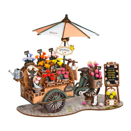 A detailed DIY kit of the Blossom Cart Model made by Robotime. It features a miniature flower cart with colorful flower pots, a large umbrella, and a chalkboard sign. The cart is decorated with various accessories like a watering can and a cute cat figure, all resting on a cobblestone base.