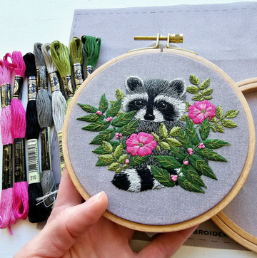 A detailed raccoon hand embroidery design in a wooden hoop, featuring a raccoon among green leaves and colorful flowers. Various embroidery threads in shades of pink, green, gray, and black are displayed beside the hoop on a light gray background.