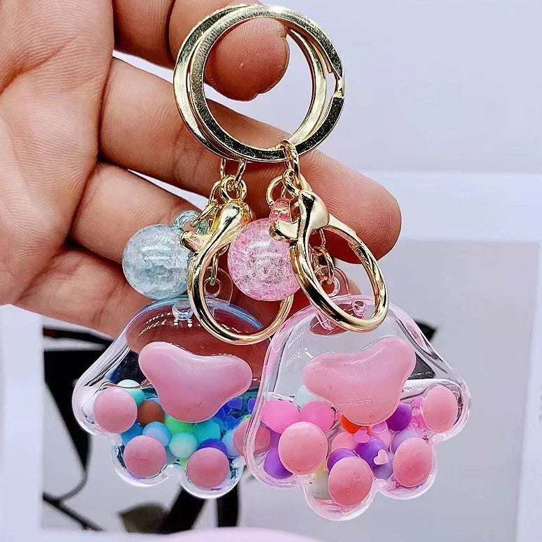 A hand holding a set of Paw Floaty Key Charms by BC Mini, featuring colorful, transparent charms shaped like paws with various soft, colorful beads inside and gold-colored hardware.