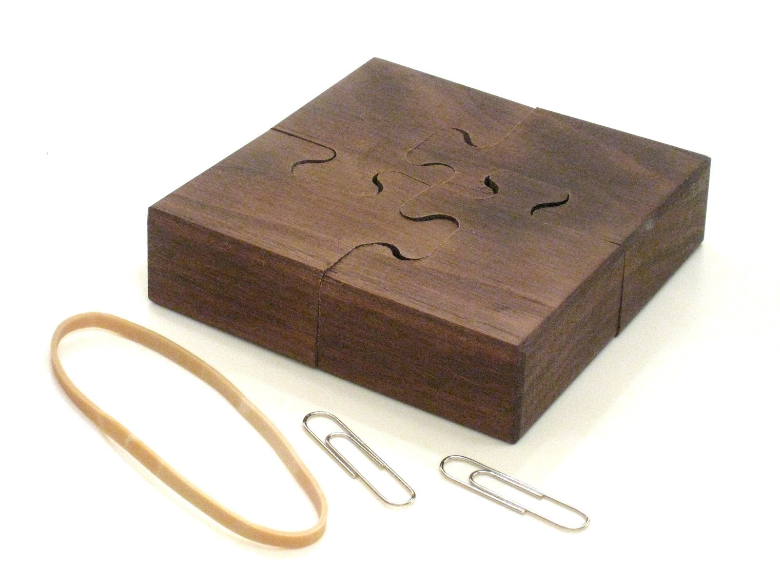 A wooden mechanical puzzle titled 'Wedge' consisting of four hinged pieces that fit together like a jigsaw. The puzzle has cut-out shapes on its surface and is accompanied by a rubber band and paperclips, showcasing its unique design and dual function as a brain teaser and decorative item.